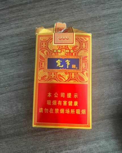 Jiaozi Kuanzhai Wuliang Mid cigarette packaging, front of a red and gold pack standing on a dark tabletop